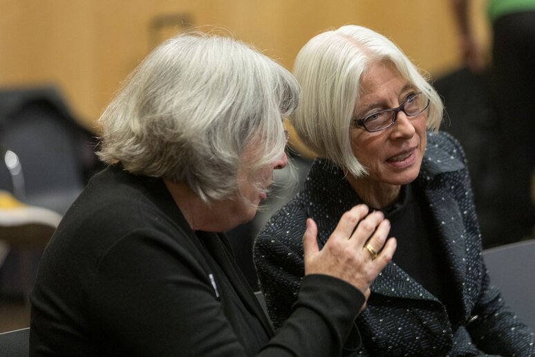 Zwei ältere Frauen sprechen miteinander. Beide haben kinnlange graue Haare. Die eine Frau neigt sich in Richtung der anderen Frau.