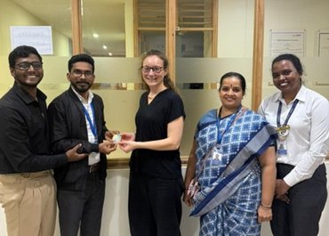 Fünf Personen stehen zusammen in einem Innenraum. Eine Frau in der Mitte hält ein kleines Objekt, das ihr von einer Person neben ihr überreicht wird.