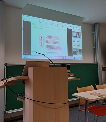 Ein Beamer projiziert die Vortragsfolien und das Bild der Referentin an die Wand.