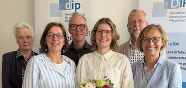 Sechs Personen stehen gemeinsam vor einer DIP-Roll-up-Wand und schauen in die Kamera. In der Mitte hält eine Frau einen Blumenstrauß, die Gruppe wirkt freundlich und lächelt; alle sind in heller, gepflegter Kleidung gekleidet.