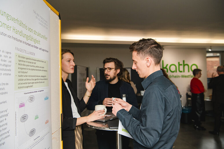 Drei Teilnehmende stehen an einem Tisch und diskutieren. Links von ihnen ist eine Stellwand mit einem Forschungsplakat.