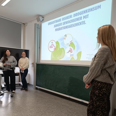 Eine PPT-Folie wird an die Wand geworfen; drei Studierende stehen rechts und links des Bildes.