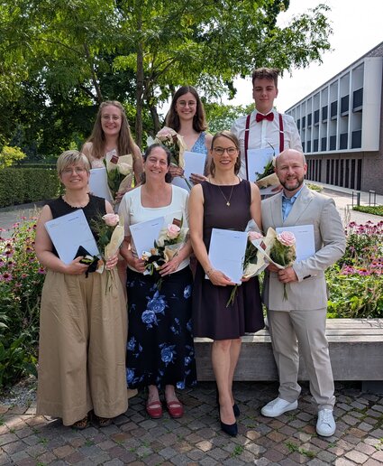 Die Absolvent_innen stehen mit ihrem Zeugnis unter einem Baum und lächeln in die Kamera.