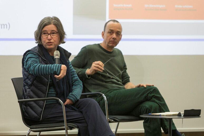 Eine Frau mit Behinderung sitzt auf der Bühne und spricht. Sie hat eine extra Halterung für das Mikrofon weil sie es nicht greifen kann. Neben ihr sitzt ein Mann.