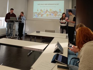 1. Folie der PPT wird an die Wand geworfen; vier Studierende stehen rechts und links des Bildes.