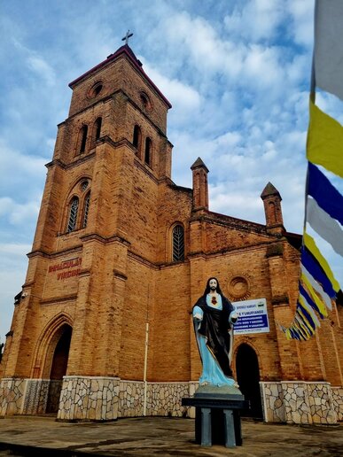 eine Kirche in hellbrauner Aussenfassade vor blauem Himmel. Vor der Kirche steht eine Jesus-Statue, an der Seite zur Kirche hängen Wimpel in den Farben Weiß, Blau und Gelb.