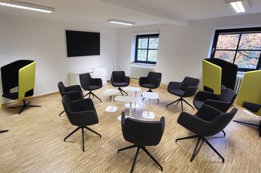 Auf dem Bild wird der neu ausgestatte Multifunktionsraum des Standorts Aachen mit einer Sitzgruppe und einem Monitor in einem modern wirkenden Raum.
