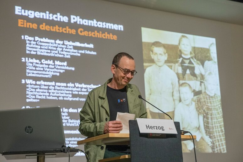 Ein Mann steht hinter einen Rednerpult. Im Hintergrund eine läuft eine Präsentation zu "Eugenischen Phantasmen- eine deutsche Geschichte"