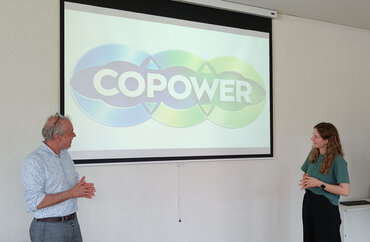 Zwei Personen stehen vor einer Leinwand mit dem farbigen CoPoweR-Logo während einer Präsentation oder eines Workshops.  Der Mann links trägt ein gemustertes Hemd, die Frau rechts ein grünes T-Shirt, und beide wirken engagiert und interaktiv im Rahmen des Projekts.