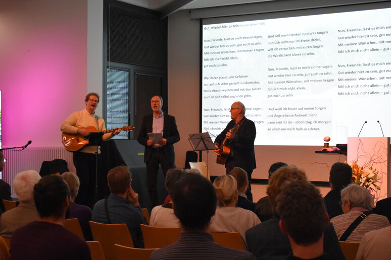 Der Musiker und Christof Stock stehen mit Gitarre auf der Bühne, dazwischen Jörg Baur