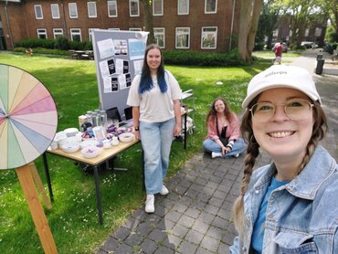 Ein Informationsstand auf der Wiese mit einem Tisch, einem "Glücksrad" und einer Person im Vordergrund