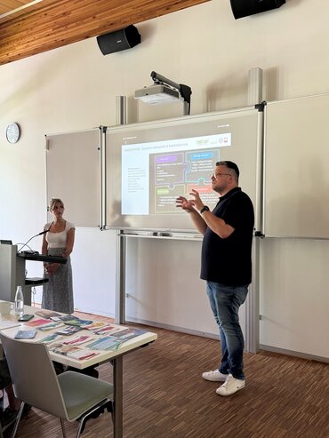 Ein Mann und eine Frau stehen vor einer Präsentation. Der Mann spricht und gestikuliert.