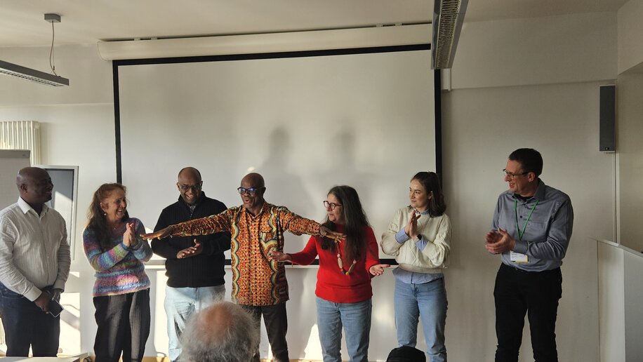 Sieben Personen stehen in einer Reihe vor einer Leinwand und klatschen. Einer trägt ein buntes Hemd und gestikuliert mit ausgebreiteten Armen. Die anderen lächeln und applaudieren. Die Stimmung ist lebhaft und positiv. Der Raum ist hell erleuchtet und wirkt freundlich und offen.