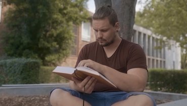 Ein Student sitzt vor einem Baum und liest konzentriert in einem Buch.