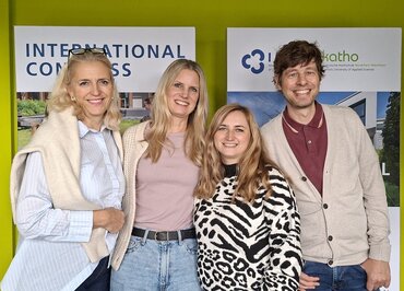 Vier Personen lachen in die Kamera. Sie halten sich in den Armen und stehen nebeneinander, vor zwei Aufstellern.