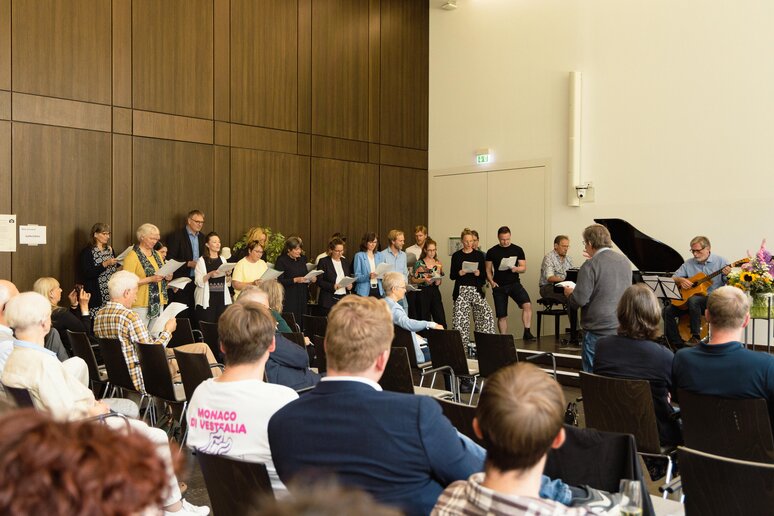 Kolleg_innen singen in einem Chor zusammen; begleitet werden sie von Klavier und Gitarre. Das Publikum hört gespannt zu.
