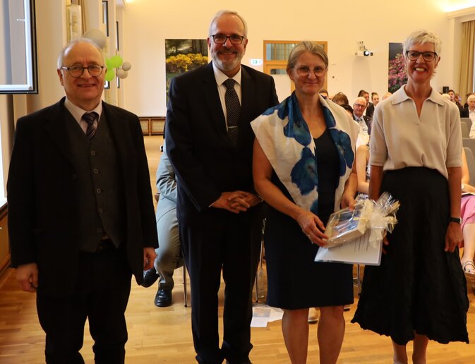 Eine Frau hält eine Urkunde und ein Geschenk in der Hand, neben ihr blicken zwei Männer und eine Frau in die Kamera.