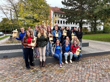 Eine Gruppe von über 20 Studierenden und Pädagog_innen auf dem Außengelände der Paderborner katho. Einige Personen halten Nistkästen aus Holz in den Händen.