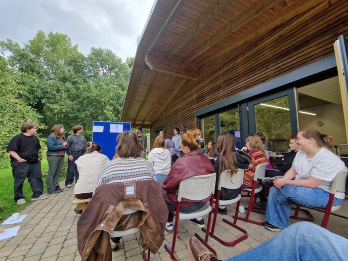 Studierende lauschen auf einer Terrasse hintereinander sitzend einem Vortrag.