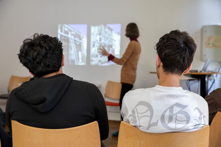Zwei Personen schauen auf einer Leinwand, wo eine Frau einen Vortrag hält.