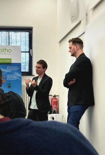 Zwei Männer, beide stehen, einer redet, einer hört zu und lehnt etwas an der Wand.