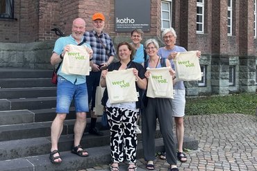 Sechs ehemalige katho-Studierende stehen vor dem Paderborner katho-Eingang. In der Hand halten sie alle einen Beutel der katho mit dem Aufdruck "wertvoll".