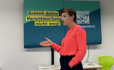 Das Foto zeigt Frau Verlinden seitlich mit Blick nach links. Sie trägt eine rote Bluse und zeigt mit ihrer rechten Hand nach vorne. Hinter ihr befindet sich ein Plakat an der Wand mit dem Schriftzug: Schieb deine Verantwortung nicht weg!