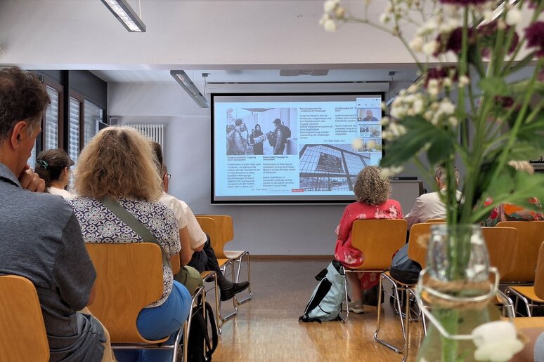 Auf einer Leinwandpräsentation ist ein Bild mit Frauen, Kindern und zwei Soldaten zusehen, ergänzt mit Texten. Menschen in Sitzreihen schauen Richtung Leinwand.