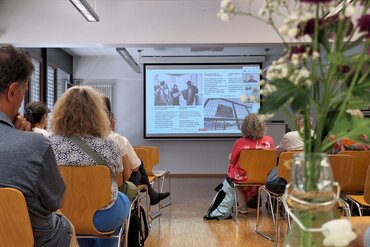 Auf einer Leinwandpräsentation ist ein Bild mit Frauen, Kindern und zwei Soldaten zusehen, ergänzt mit Texten. Menschen in Sitzreihen schauen Richtung Leinwand.
