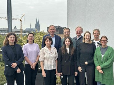10 Pesonen stehen in zwei Reihen hintereinander und lächeln freundlich in die Kamera. Im Hintergrund sind die Spitzen des Kölner Doms zu sehen.