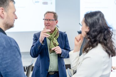 Ein Mann in einem dunklen Blazer und grünem Schal steht im Gespräch mit einer Frau und einem weiteren Mann. Im Kontext handelt es sich um Prof. Dr. Patrick Isele oder Prof. Dr. Michael Obermaier im Gespräch mit Dr.in Irina Wolf, der Leiterin des Instituts für Sozialarbeit und Sozialpädagogik (ISS) Frankfurt.