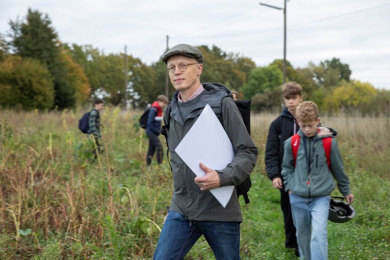 Ein Mann läuft auf einer Wiese mit einer laminierten Karte in der Hand. Ihm folgen einige Schüler:innen.