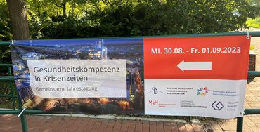 Das Foto zeigt die Plakate des Jahreskongress der Deutschen Gesellschaft für Sozialmedizin und Prävention auf denen ein Pfeil nach Links als Wegweiser zeigt.
