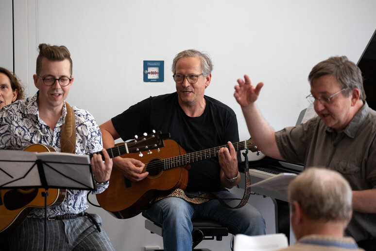 Die musikalische Begleitung übernimmt ein Gitarrenspieler und ein Klavierspieler; ein älterer Herr übernimmt das Dirigieren.