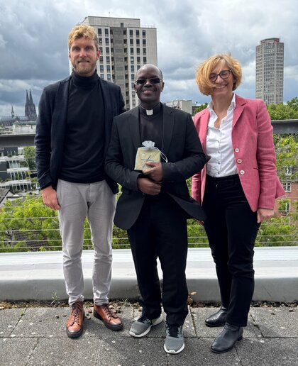 Felix Nuss, Jean Bosco Barisbeshya und Barbara Schermaier-Stöckl stehen zusammen. Im Hintergrund sind zwei Hochhäuser und der Kölner Dom.