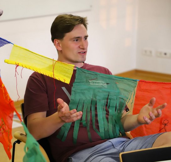 Ein Student sitzt vor einer Schnur mit gelben, roten und grünen Flaggen. Er spricht und gestikuliert.