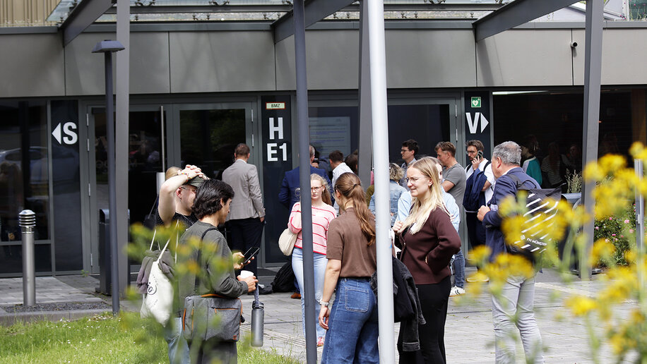 Mehrere Menschen stehen in kleinen Gruppen im Außenbereich eines Hochschulgebäudes.