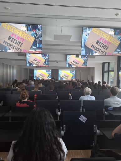Zahlreiche Menschen sitzen in Reihen in einem Vorlesungssaal. auf den Monitoren über ihnen steht "Menschenrechte statt rechte Menschen".