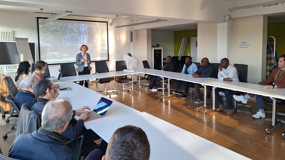 In einem Konferenzraum sitzen Menschen an einem großen, U-förmigen Tisch. Eine Frau steht präsentierend vor einer Leinwand, auf der ein Bild projiziert wird. Die Teilnehmenden hören aufmerksam zu, einige mit Laptops vor sich. Der Raum ist hell und modern gestaltet, mit viel natürlichem Licht.