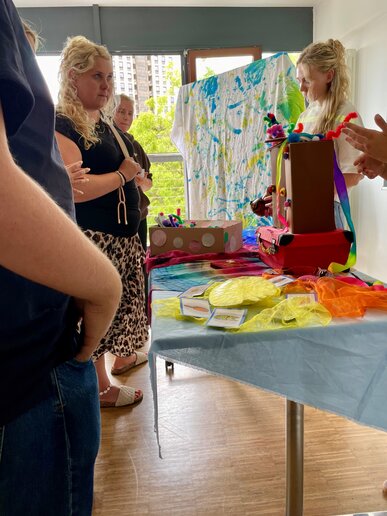 Teilnehmende und Studierende sind im interessierten Austausch über das vorgestellte Projekt. Dazwischen steht ein Tisch auf dem Materialien des Projektes stehen, beispielsweise ein selbstgebastelter Koffer.