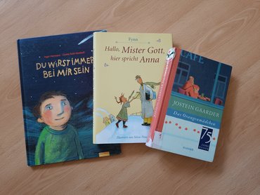 Drei Kinderbücher mit den Titeln "Du wirst immer bei mir sein", "Hallo, Mister Gott, hier spricht Anna" und "Das Orangenmädchen" liegen auf einem Tisch.