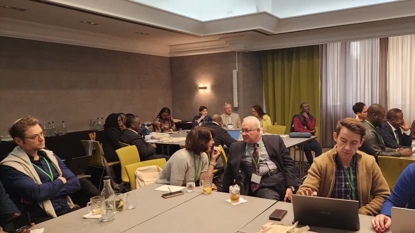 In einem Konferenzraum sitzen mehrere Personen an Tischen. Einige haben Laptops vor sich, während andere miteinander sprechen. Die Teilnehmenden wirken konzentriert und diskutieren in einer professionellen Atmosphäre. Im Hintergrund sind Vorhänge und weitere Menschen zu sehen, die ebenfalls in Gespräche vertieft sind.