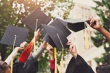 Hände werfen ihre Studentenhüte in die Luft. Im Hintergrund sind Häuser und grüne Bäume zu sehen.