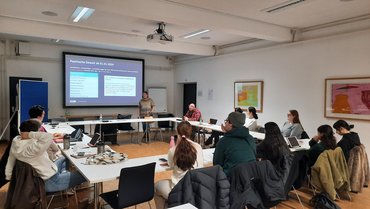 Das Bild zeigt einen gut besetzten Seminarraum, vorne steht jemand an der Leinwand und referiert.