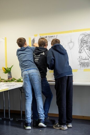 Schüler:innen malen mit Kohle auf an der Wand befestigtem Papier.
