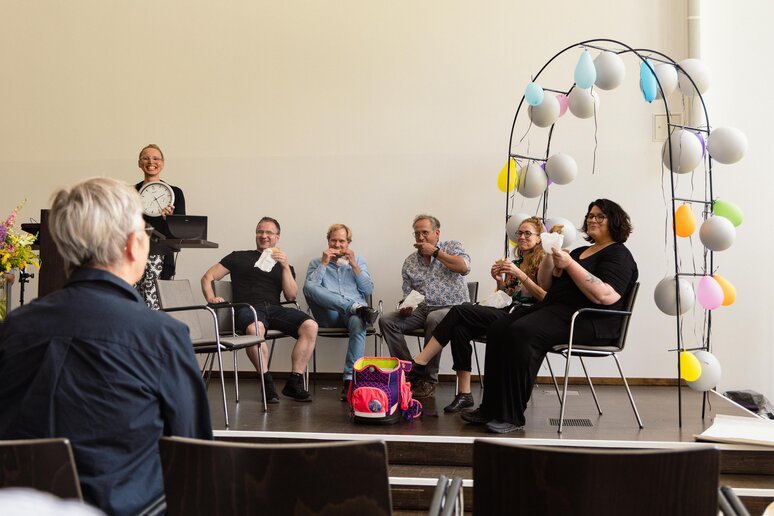 Kolleginnen und Kollegen sitzen auf der Bühne vor einem Bogen geschmückt mit Luftballons. Die geehrte Kollegin folgt interessiert dem Gespräch auf der Bühne.