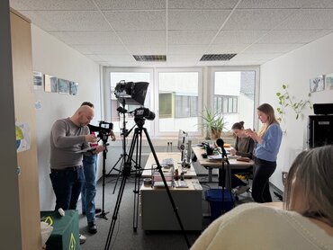 Zwei Frauen hinter einem Schreibtisch; Kameramann und Kameras stehen vor dem Schreibtisch.