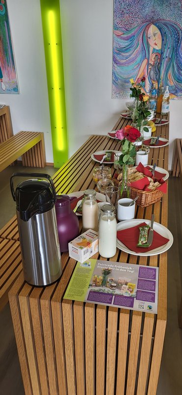 Ein länglicher Holztisch, der mit Frühstückstellern, Tassen, Milch, Kaffee etec. gedeckt ist.