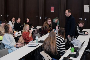 Studierende sitzen an Tischreihen im Hörsaal. Eine Studentin in der Bildmitte spricht in ein Mikrofon, rechts von ihr steht der Dozent.