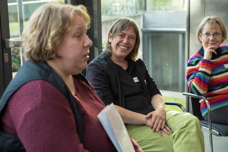 Eine Frau im Vordergrund. Sie hält Notizen in der Hand und erklärt etwas. Zwei weitere Frauen im Hintergrund die sie anschauen und zuhören.